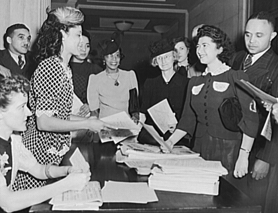 photograph shows Jewel Mazique handing papers across a table as a group of Library employees looks on