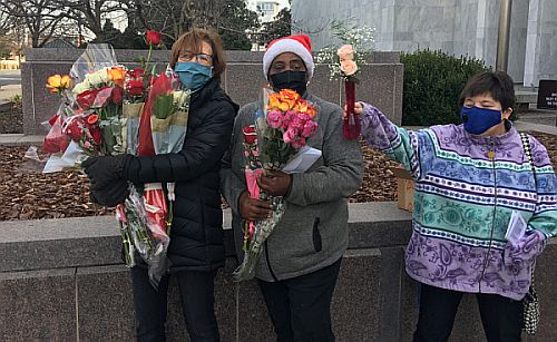 Three women outdoors wearing winter coats, holding multiple bouquets of flowers.